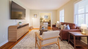 A living room with a flat-screen TV, a wooden cabinet, a brown leather sofa, a striped armchair, a small table, and a lamp. - Epic Game Room Hangout with Grill, King Beds & 5 TVs (Valdosta)