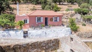 Una villa rosa con techo de tejas, un pequeño patio con muebles de exterior y vistas a una colina con árboles.