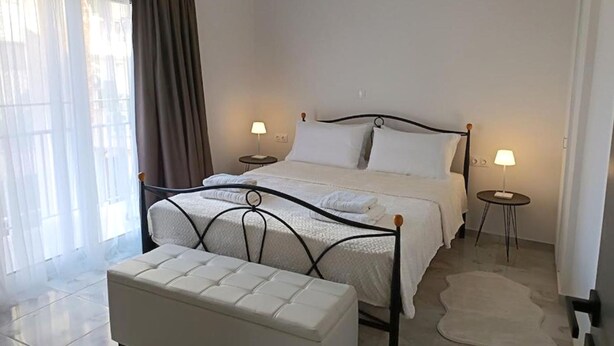 Quarto com cama, mesas de cabeceira, um banquinho e uma janela grande com cortinas.