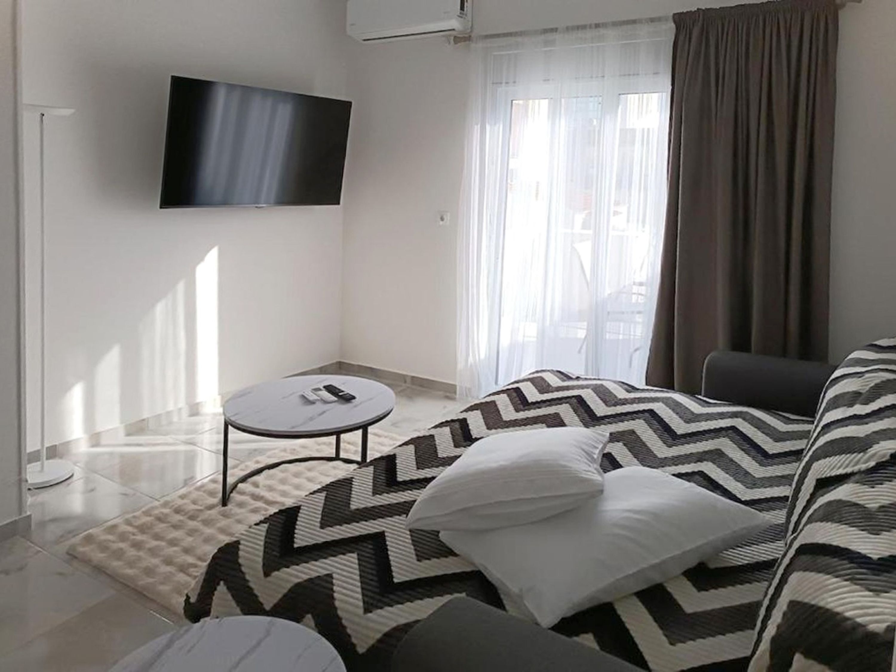 Une chambre moderne avec un lit, un téléviseur fixé au mur, une petite table ronde et une fenêtre avec des rideaux.