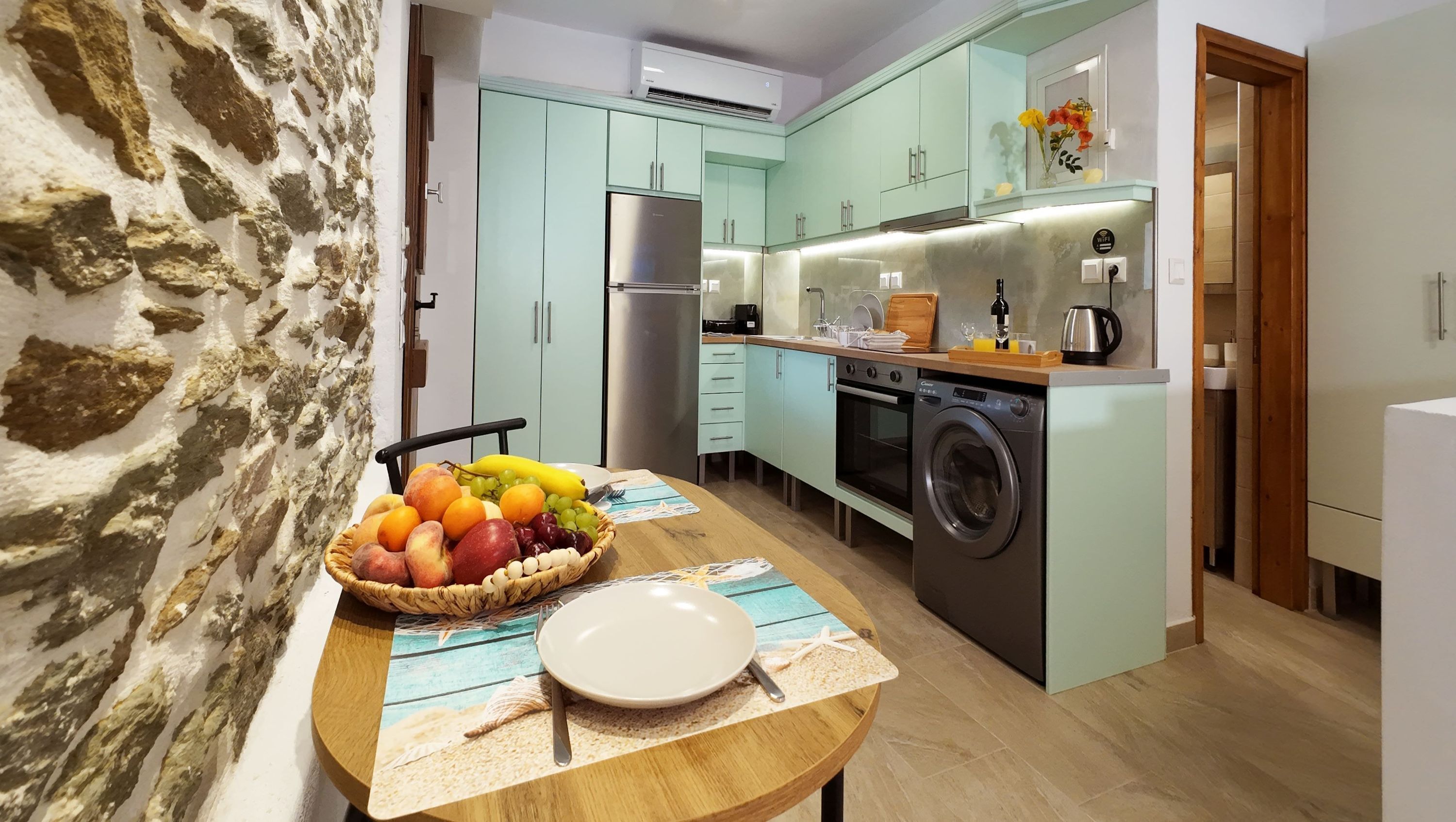 Uma cozinha compacta com armários verde-claros, máquina de lavar embutida e um espaço de refeições com uma cesta de frutas e um jogo americano com tema de praia.