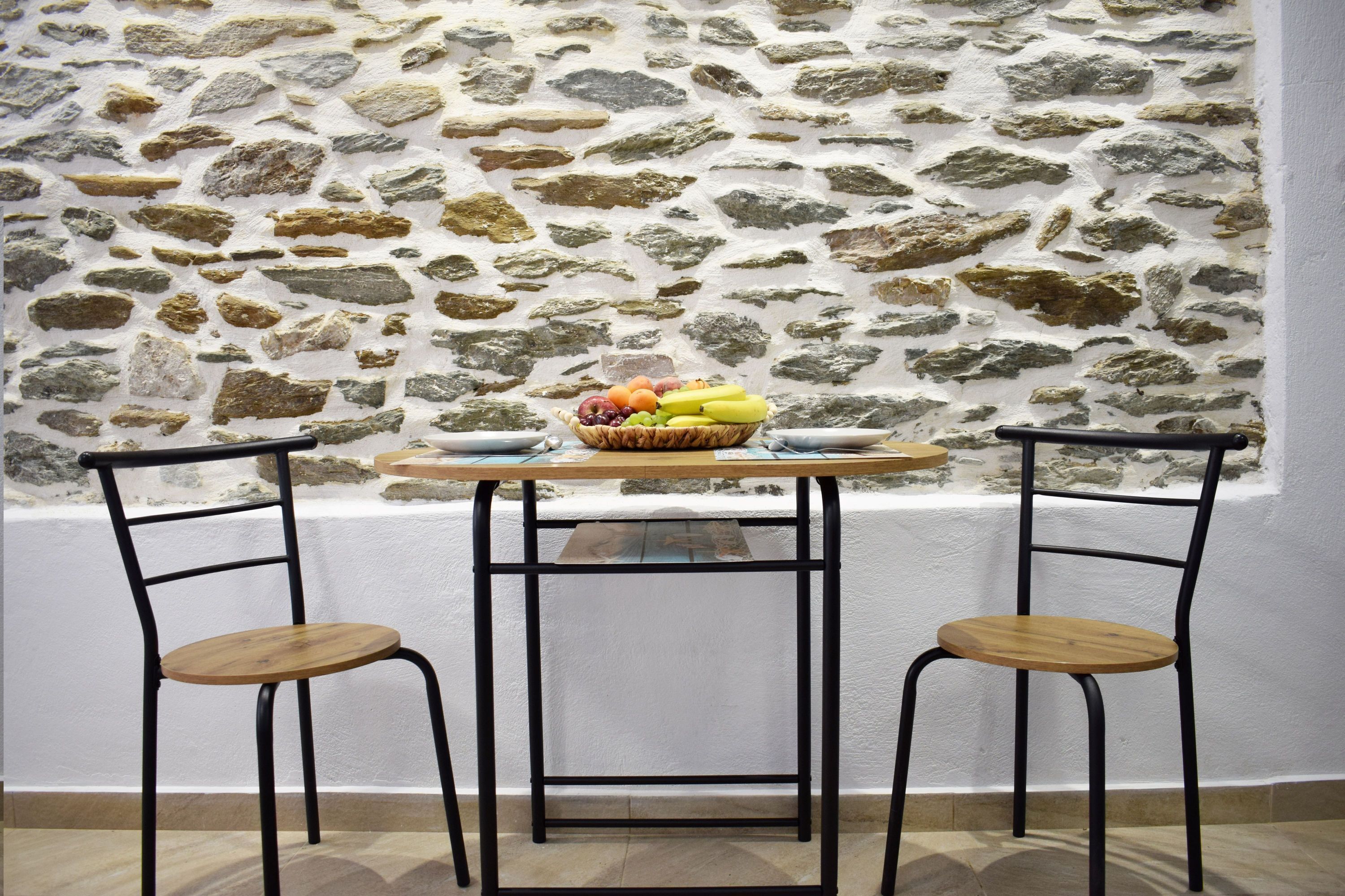 A room with a stone wall, a round wooden table, two metal chairs, and a fruit basket on the table.