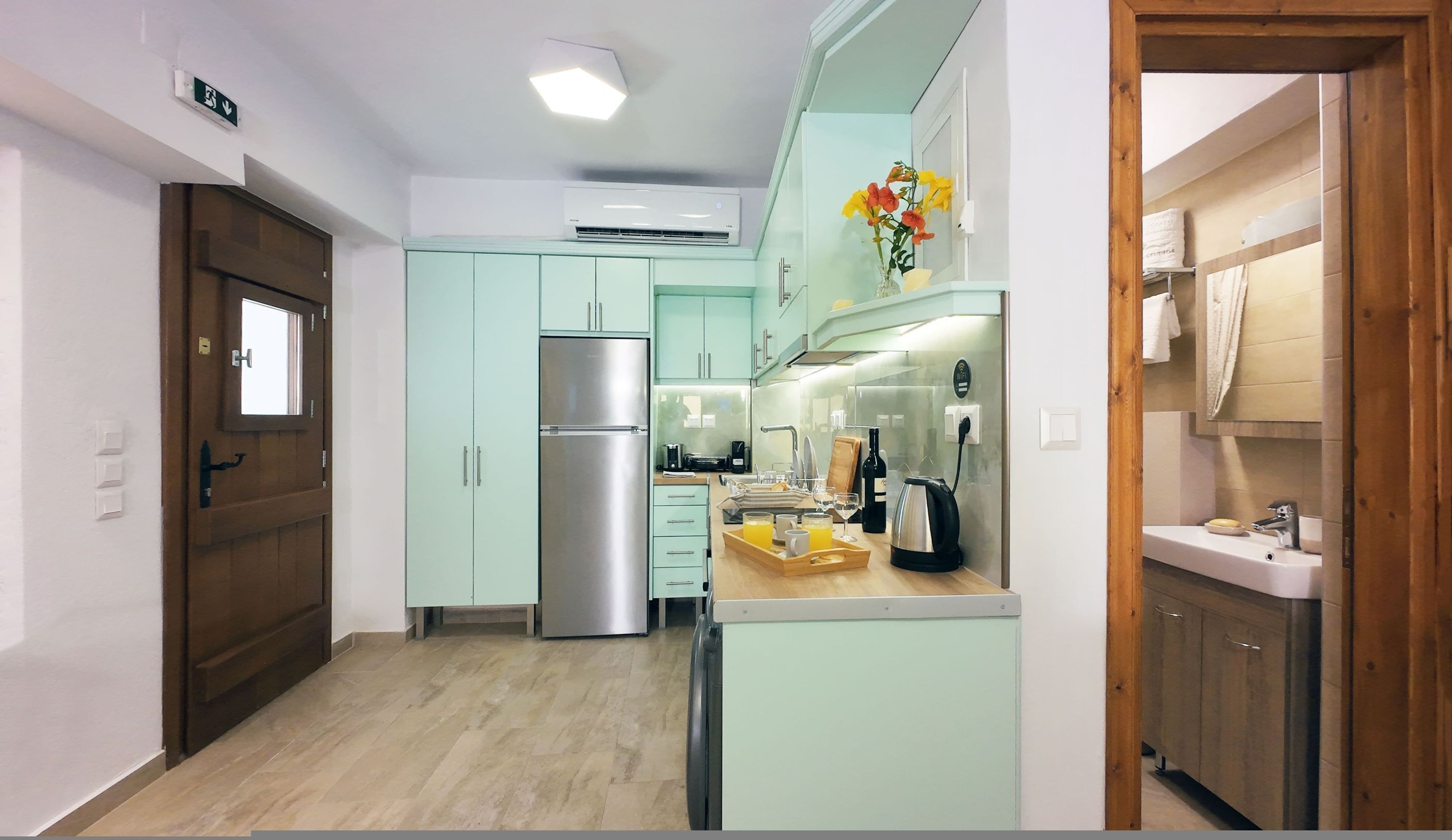 Cozinha moderna com armários verde-clarinho, geladeira de aço inoxidável e bancada de madeira com uma chaleira e uma bandeja de suco de laranja.