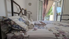 Un dormitorio con cama, ropa de cama floral, cabecera de madera, ventana con cortinas y balcón con barandilla metálica.