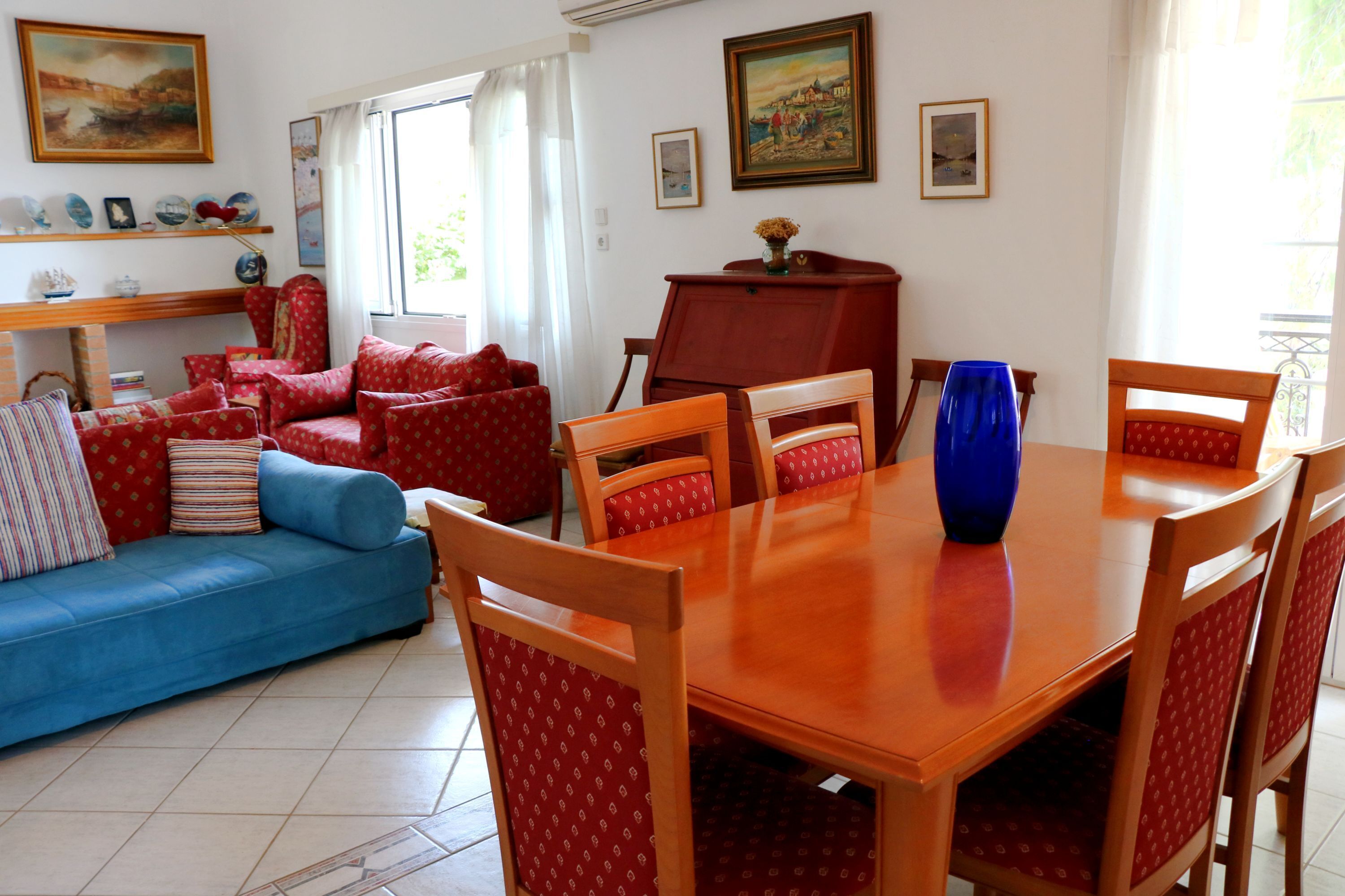 Un salon avec un canapé bleu, des fauteuils à motifs rouges, une table à manger en bois et un vase bleu.