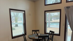 A dining area with a round table and four chairs, large windows with blinds, and a view of outdoor seating.