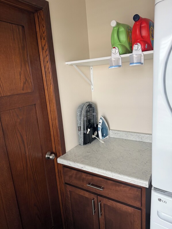 Una cocina con puerta de madera, encimera, microondas empotrado, estante con botellas de detergente y un planchado de pared.