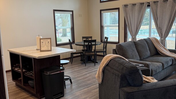 A spacious living area with a dark gray sofa, a dining table with chairs, and a kitchen area with a countertop and cabinets.