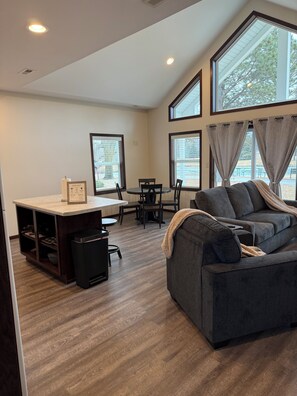 A spacious living area with a dark gray sofa, a dining table with chairs, and a kitchen area with a countertop and cabinets. - Cozy 1-bedroom house in beautiful Brookings on the walking trail (Brookings)