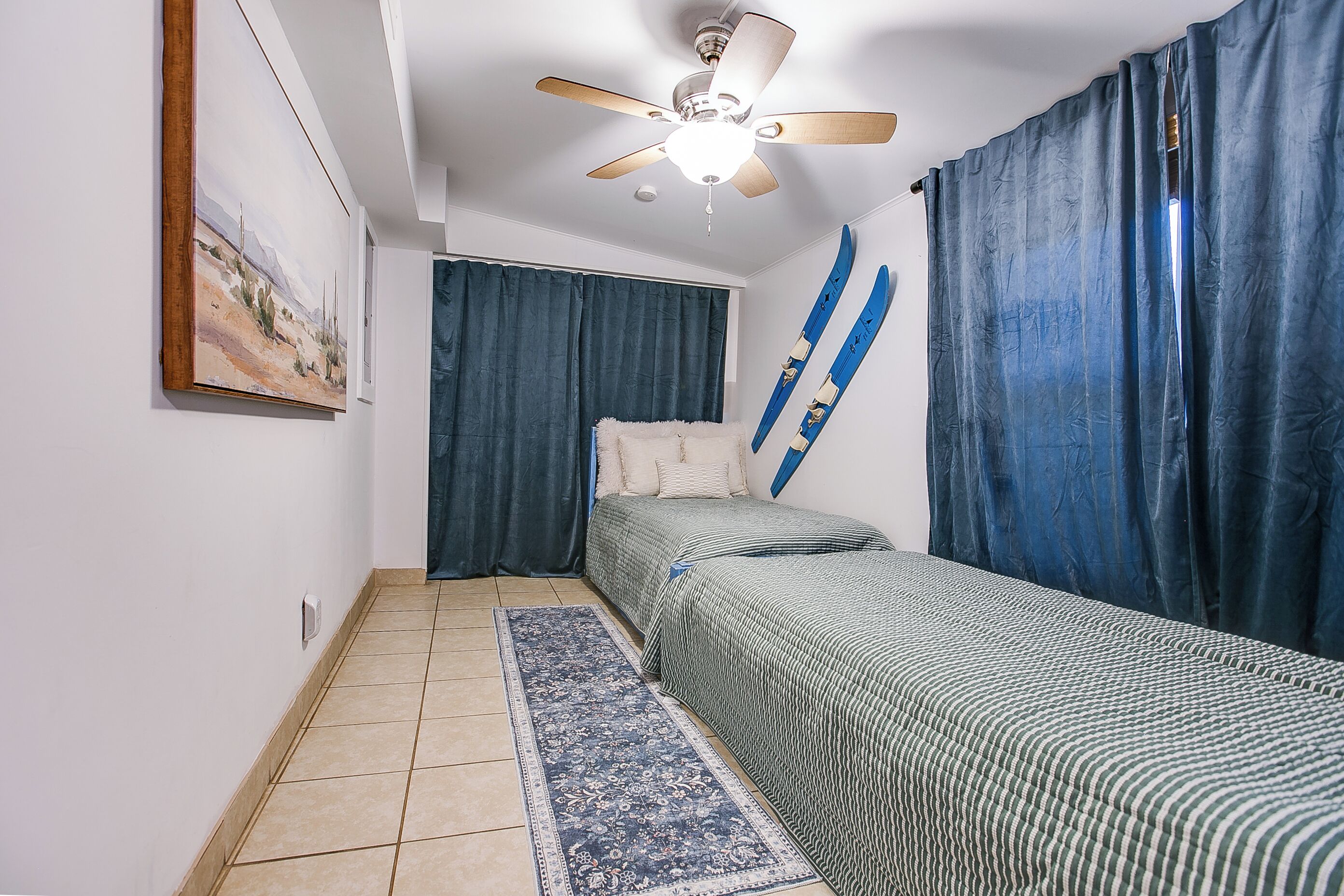 Une chambre à coucher avec un lit, un ventilateur de plafond, un tableau au mur et une fenêtre avec des rideaux.