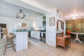 Cocina moderna con armarios blancos, una isla central y electrodomésticos de acero inoxidable. Hay una zona de comedor con una mesa y sillas, y una pequeña planta decorativa sobre la mesa.