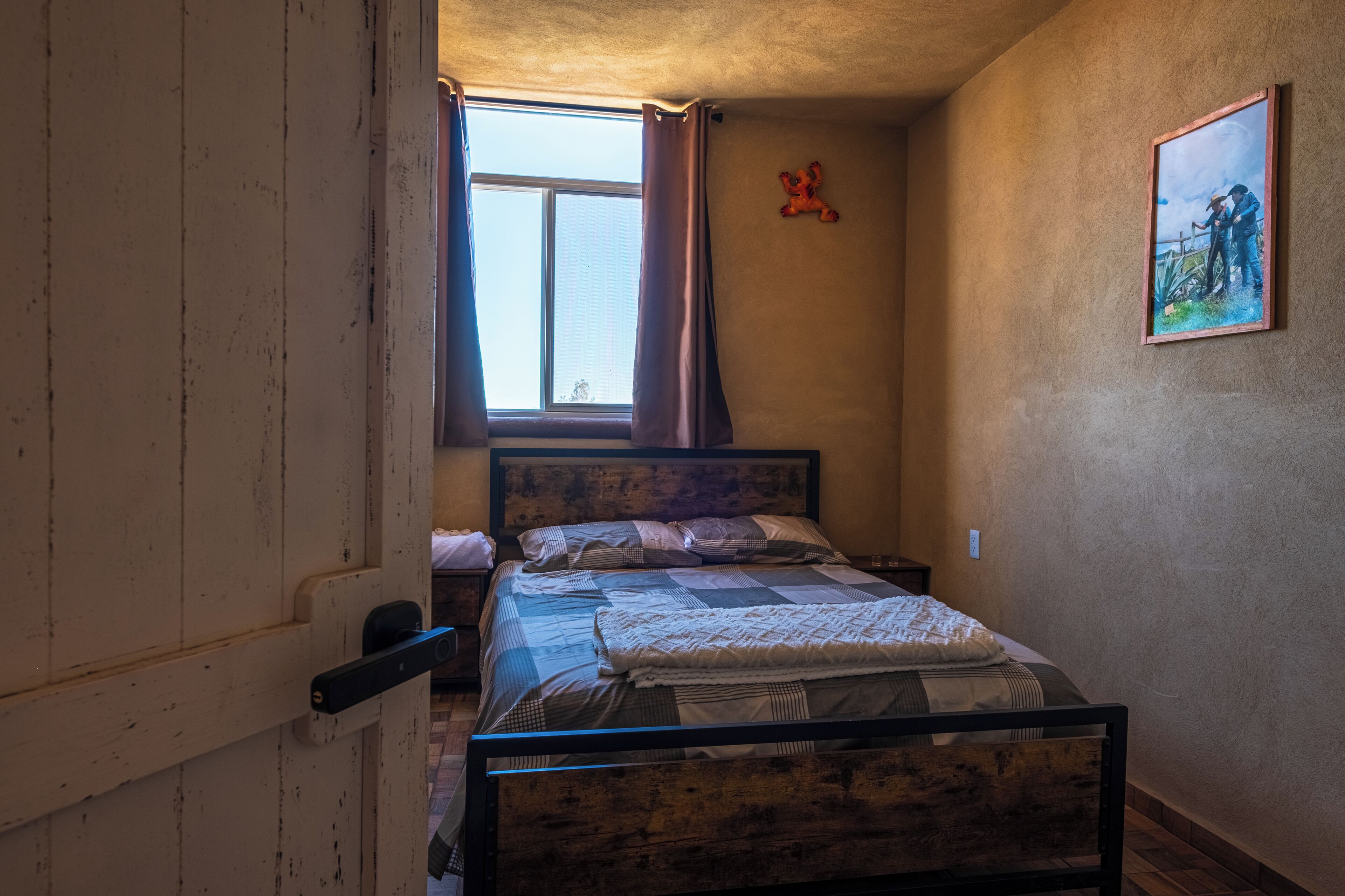Un dormitorio con una cama de madera, una ventana con cortinas, un peluche decorativo y un cuadro en la pared.