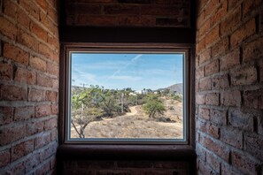 A window with a view of a dry, hilly landscape with sparse vegetation and a few buildings in the distance. - Cuanax Hotel (Pozos)