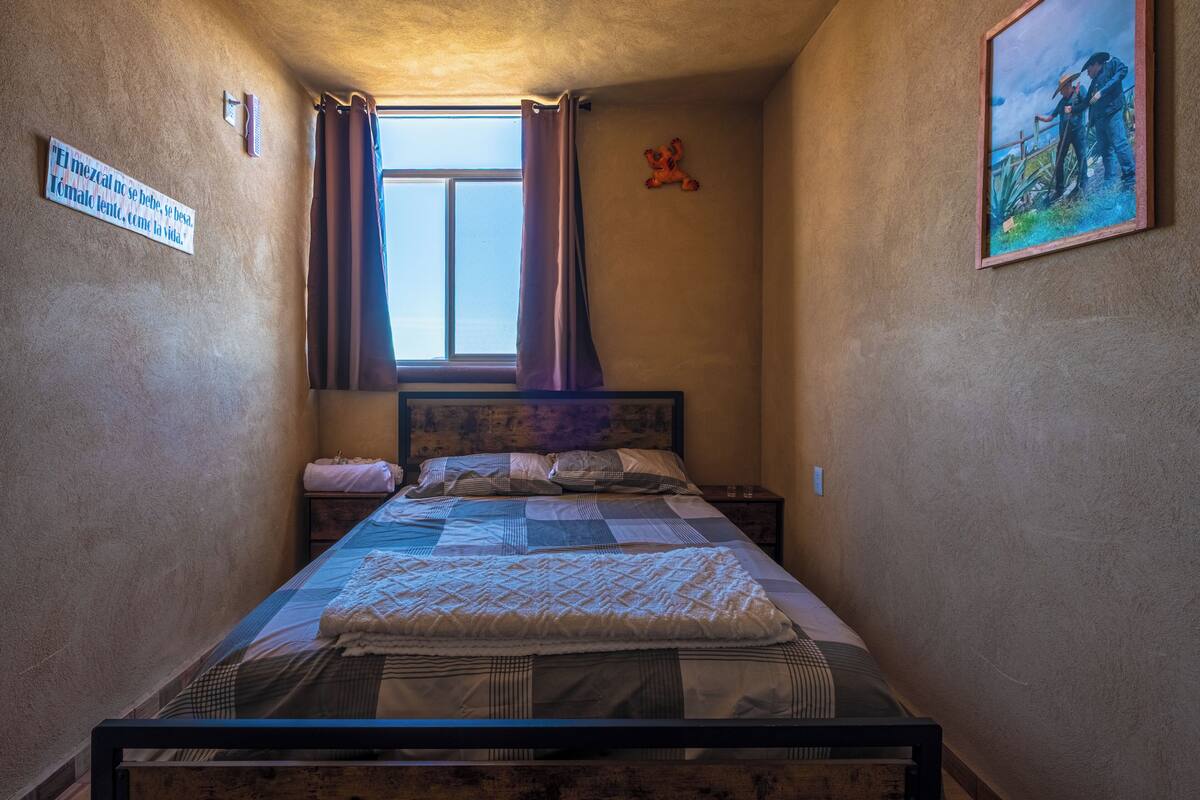 A narrow room with a bed, a window with curtains, a wall-mounted picture, and a sign with text.