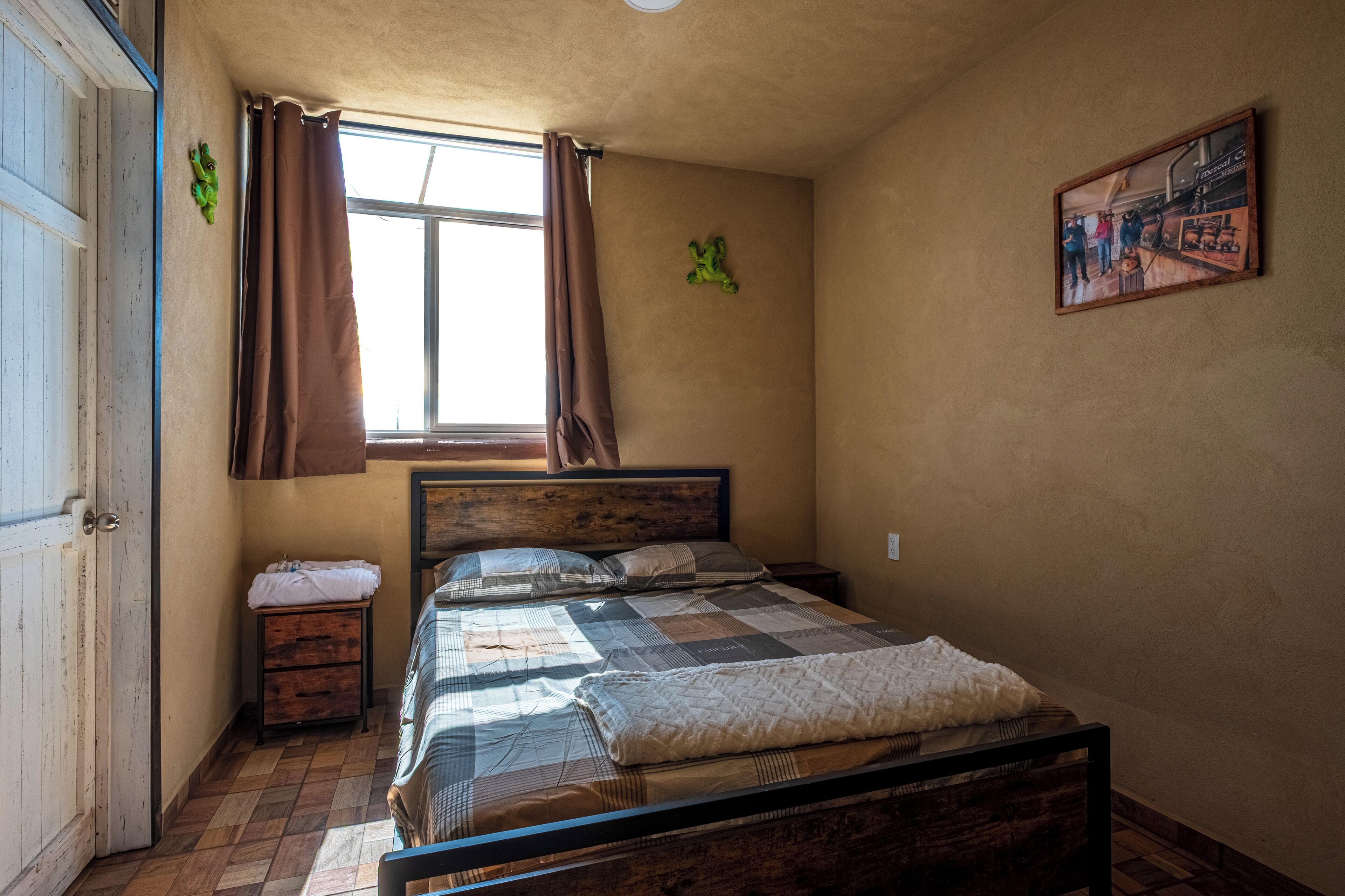 Une chambre avec un lit en bois, une petite table de chevet, une fenêtre avec des rideaux et un tableau accroché au mur.