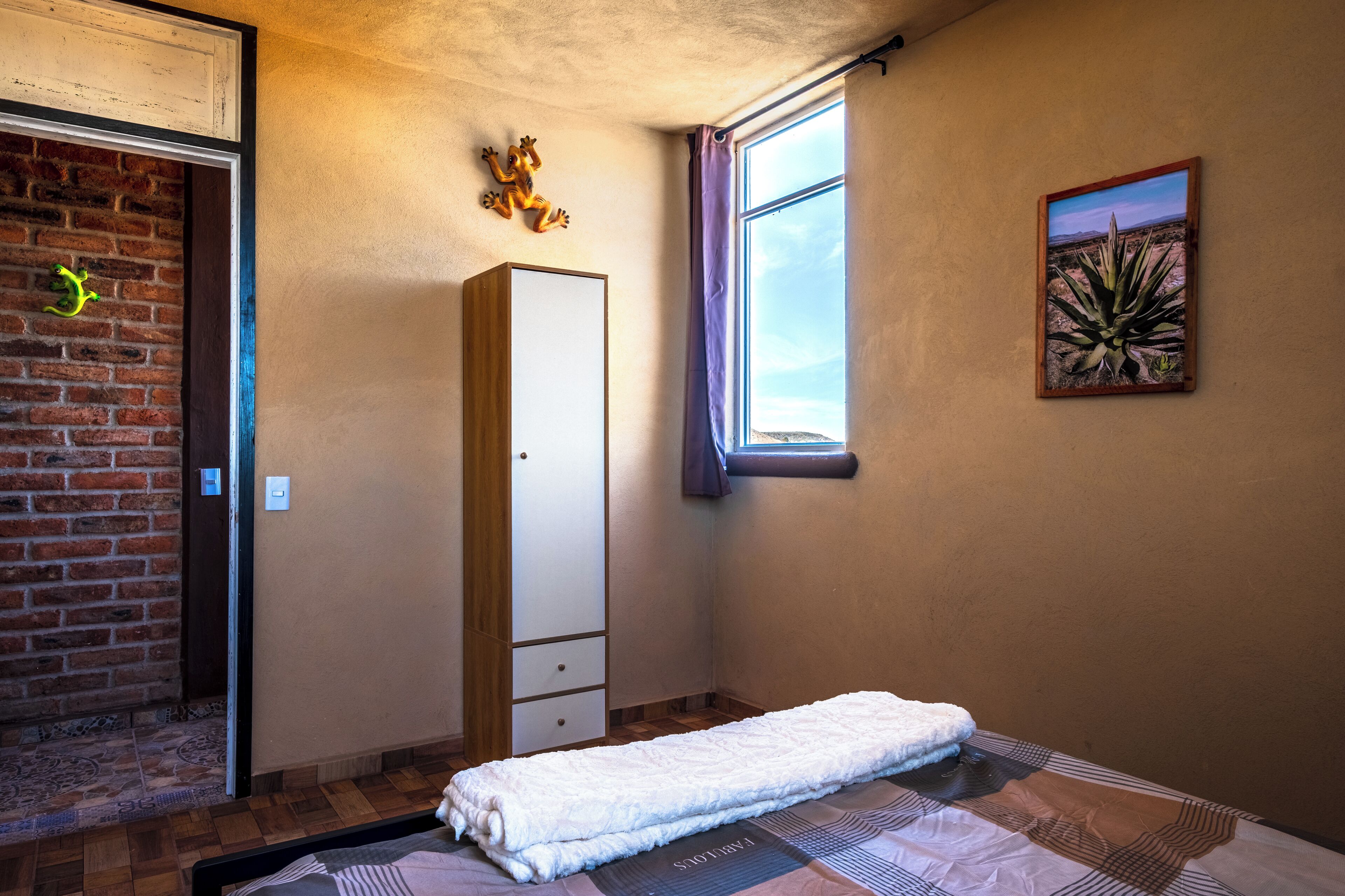 A bedroom with a bed, a wardrobe, a window with curtains, a brick wall, and a painting.