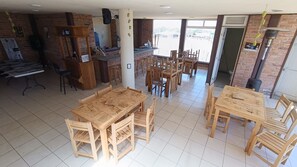 Un espacio interior amplio con varias mesas y sillas de madera, una zona de bar con taburetes y un ventanal con vistas al exterior.