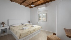 Et soveværelse med træloft, en seng med hvide sengetøj, et natbord med en lampe og et vindue med udsigt til træer.