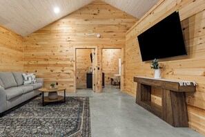 Interior de uma cabana de troncos com sofá, mesa de centro, tapete, lareira e televisão na parede.