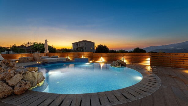 Una piscina con cubierta de madera, rodeada de rocas, y una vista al atardecer de fondo.