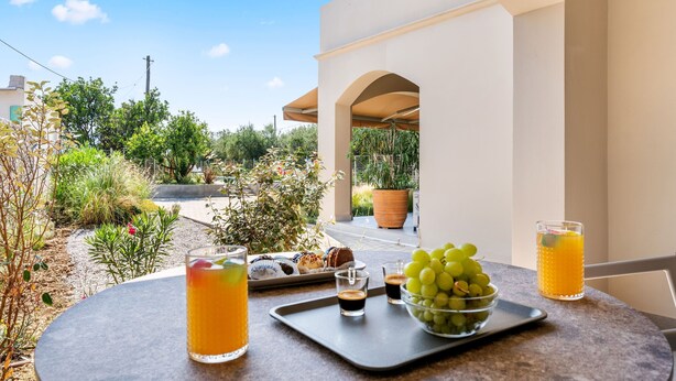 A table with a tray of pastries, a bowl of grapes, and glasses of orange juice on a balcony overlooking a garden. - garden & pool view Ground floor (Nikiana)