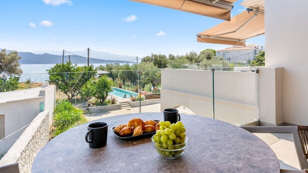 A balcony with a table set with croissants, grapes, and two black mugs, overlooking a pool and mountains. - Sea View Suite  First Floor (Nikiana)