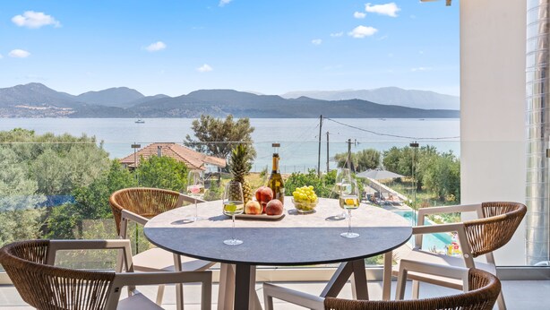 Un balcón con una mesa dispuesta para dos personas, que incluye frutas, una botella de vino y copas. La vista muestra una masa de agua y montañas distantes.