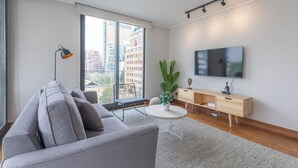 Un salon moderne avec un canapé gris, une table basse ronde, un téléviseur fixé au mur et une grande fenêtre donnant sur des immeubles.