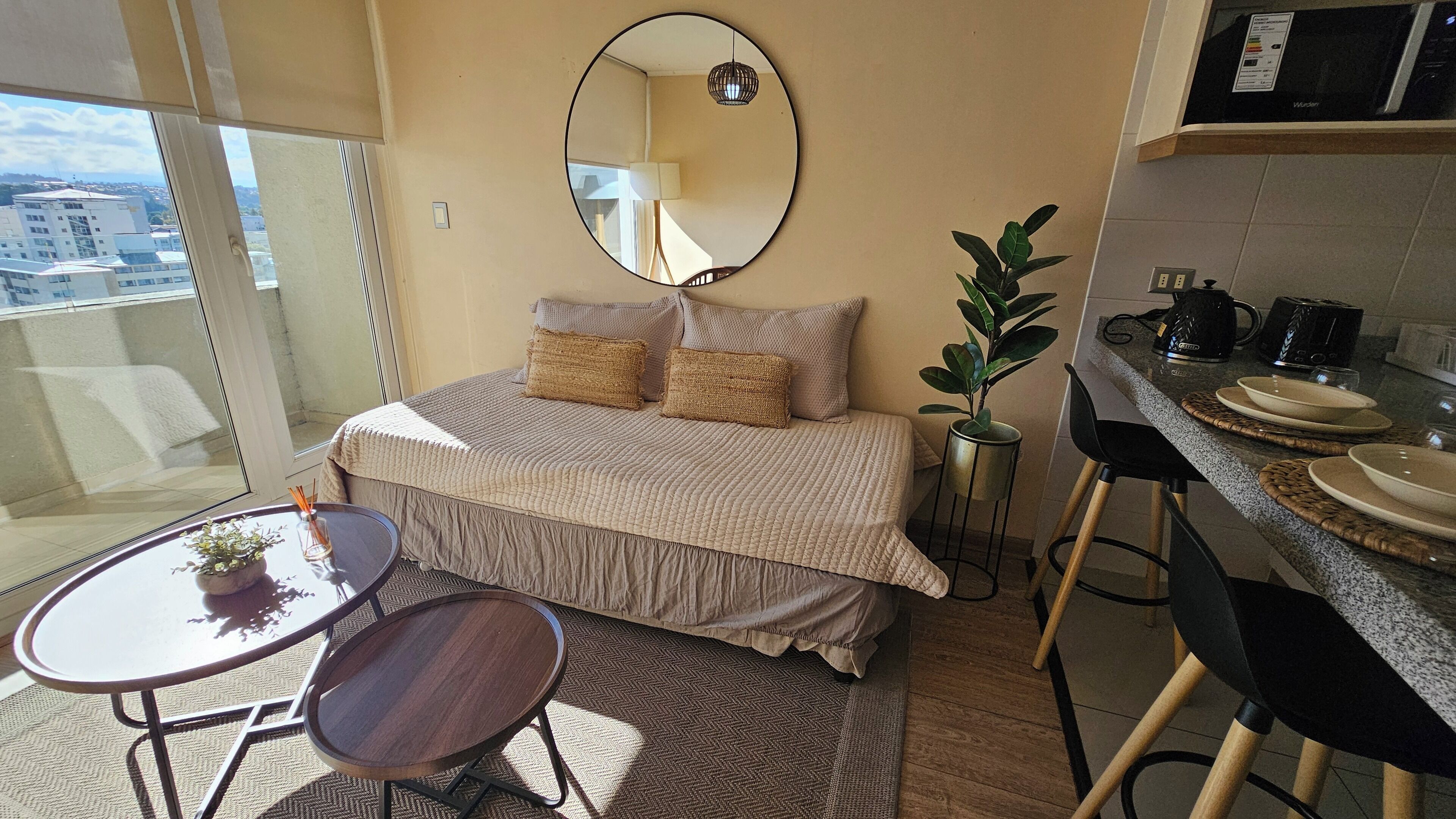 Une chambre avec un lit, un miroir rond, une petite table avec une chaise, une plante en pot et un coin cuisine.
