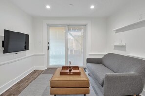 Een moderne woonkamer met een grijze bank, een houten poef, een flatscreen televisie aan de muur en een glazen schuifdeur met jaloezieën.