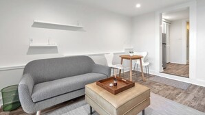 A modern living room with a grey sofa, a small round wooden coffee table, and a white chair. There is a small white shelf on the wall and a door leading to a kitchen. - The Ocean's Edge Studio (SF)