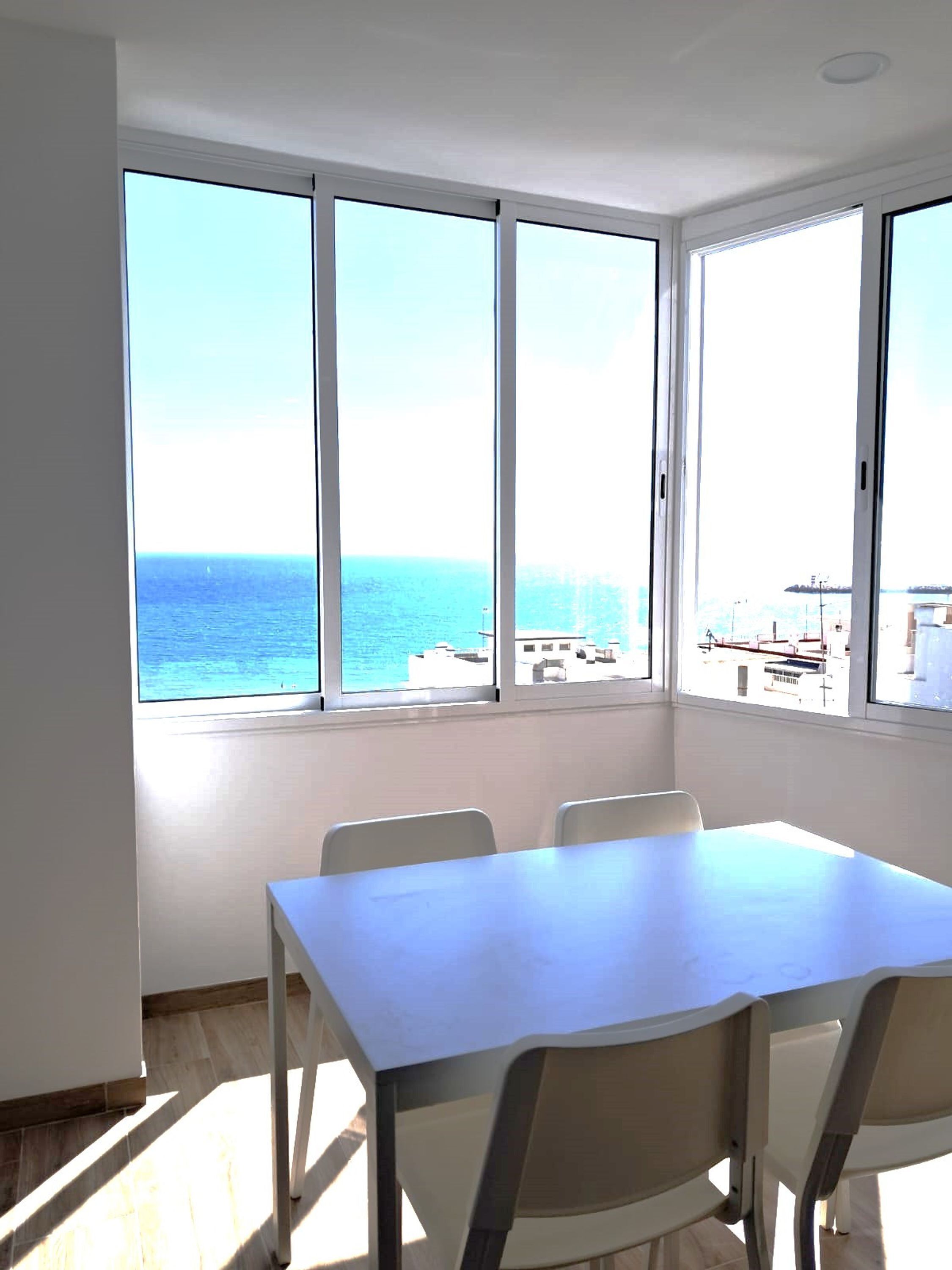 A room with a large window overlooking the sea, a white table, and four chairs.