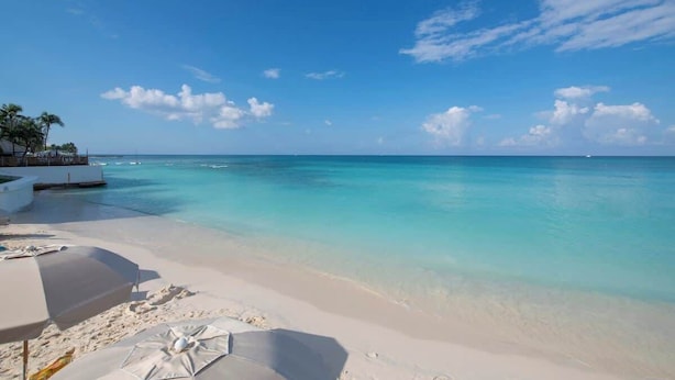 Ein Strand mit klarem türkisblauem Wasser, weißem Sand und einigen Sonnenschirmen.