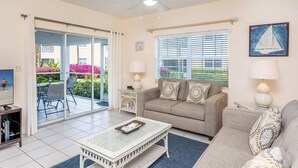 Ein Wohnzimmer mit einer Couch, einem Couchtisch und einem Fernseher. Eine Schiebetür führt zu einem Außenbereich mit Pflanzen und Blick auf den Strand.