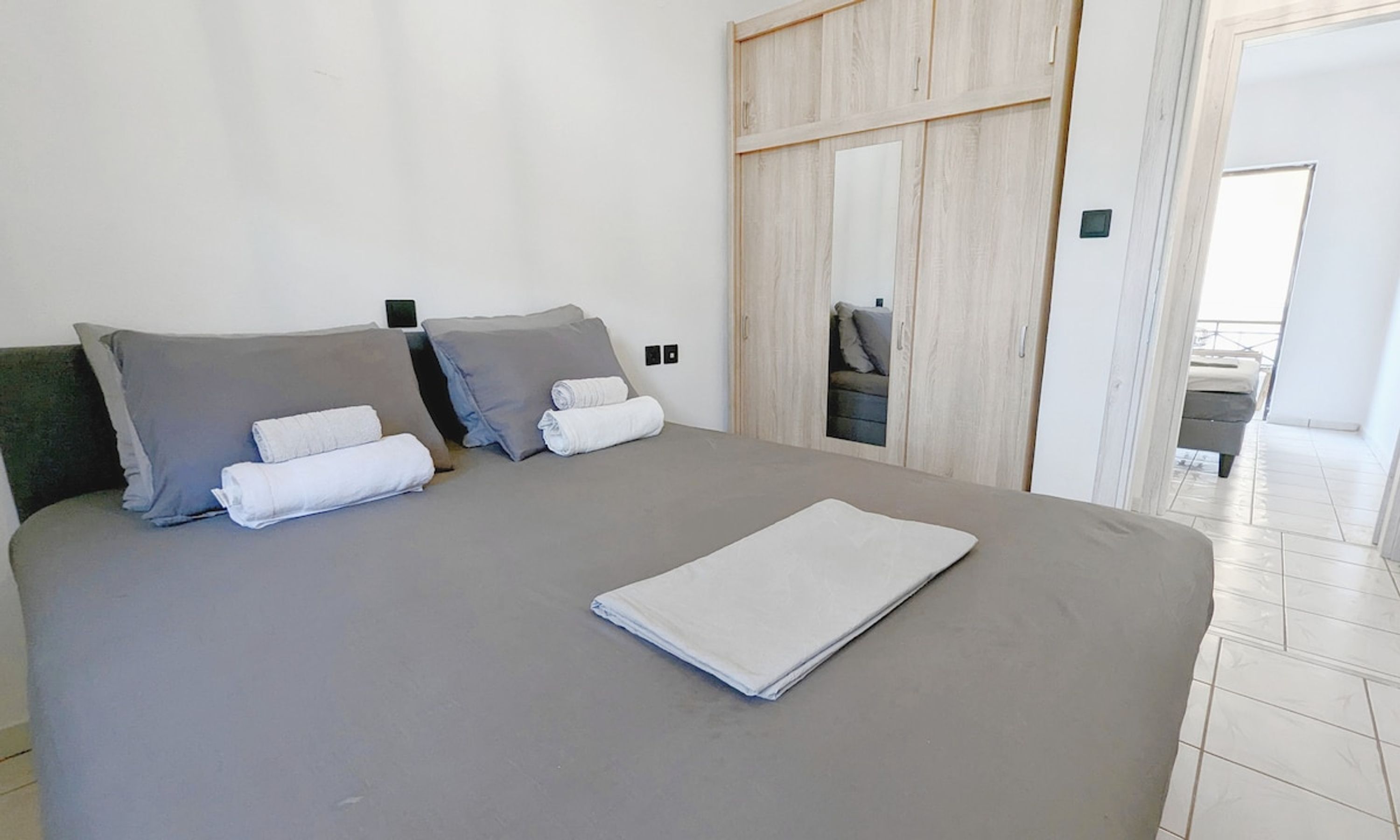 A neatly made bed with grey bedding and white towels, a wooden wardrobe, and a view of a balcony with a grey sofa.