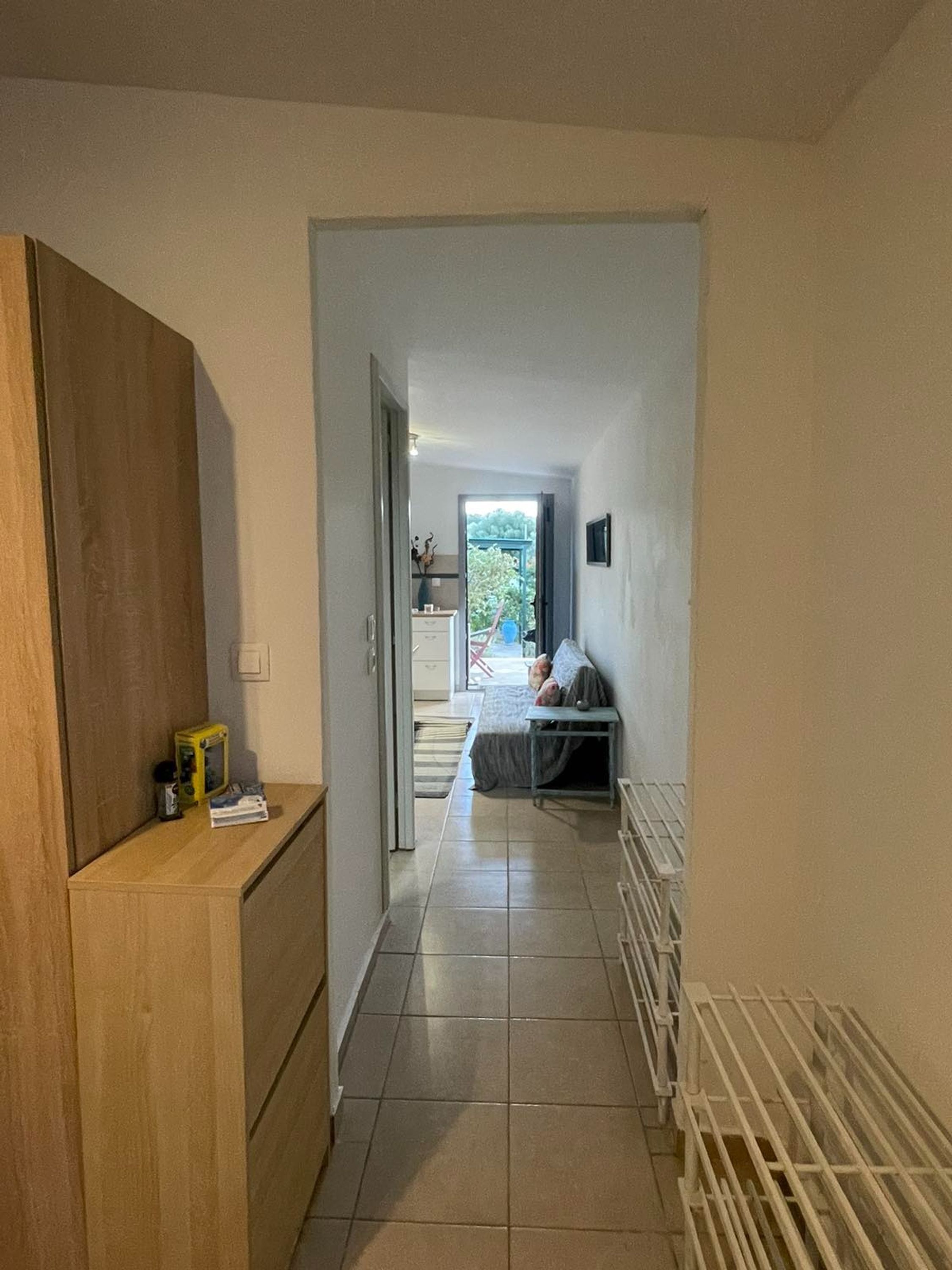 A hallway leading to a bedroom with a bed, a small table, and a chair. There is a radiator on the right and a wooden cabinet on the left.
