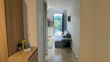 A hallway leading to a bedroom with a bed, a small table, and a chair. There is a radiator on the right and a wooden cabinet on the left.