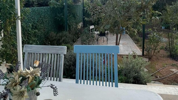 A covered patio with two white chairs, a table with a vase of flowers, and a view of a garden with a pergola and trees. - Simple Stay  Vari's House (Vari)
