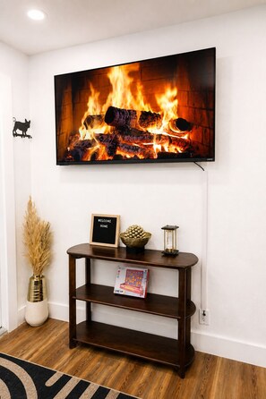 Ein an der Wand befestigter Fernseher zeigt ein Kaminfeuer, ein dunkles Holzkonsole mit einem Willkommensschild und eine Vase mit Trockenpflanzen.