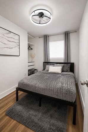 Ein Schlafzimmer mit Bett, Nachttisch, Pendelleuchte, einem Fenster mit Vorhängen und einem an der Wand hängenden Kunstwerk.
