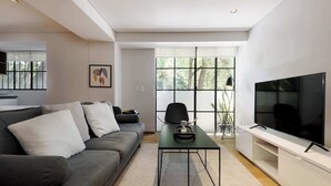 Un salon moderne avec un canapé gris, une table basse, un téléviseur posé sur un meuble et une grande fenêtre donnant sur des arbres.