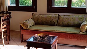 Ein Zimmer mit einer Couch, einem Holztisch und einem Kronleuchter. Zwei Fenster bieten Blick auf Grünflächen.