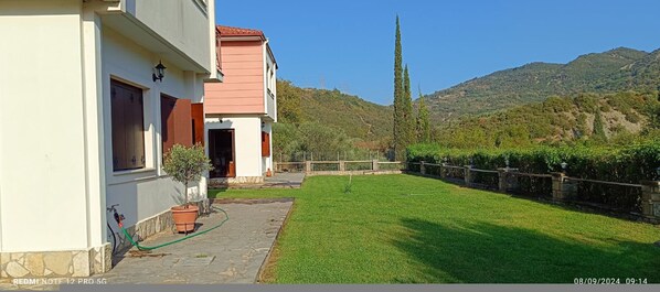Una casa con un patio empedrado y un césped bien cuidado, rodeada por una cerca de piedra y un paisaje montañoso.