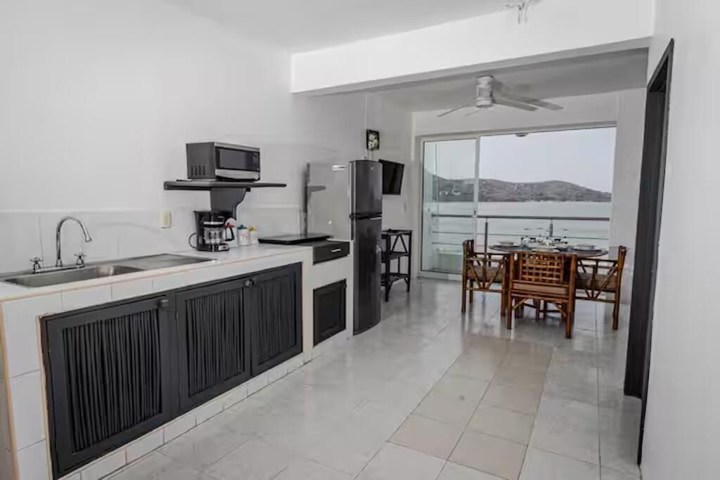 A modern kitchen with a built-in microwave, refrigerator, and sink. There is a dining area with a table and chairs, and a balcony with a view.