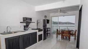 A modern kitchen with a built-in microwave, refrigerator, and sink. There is a dining area with a table and chairs, and a balcony with a view. - Beachfront Olas Bungalow @Aqua at La Madera Beach. (Zihuatanejo)