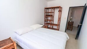 A bedroom with a bed, a wooden shelf unit, a small wooden table, and a door leading to another room. - Beachfront Olas Bungalow @Aqua at La Madera Beach. (Zihuatanejo)