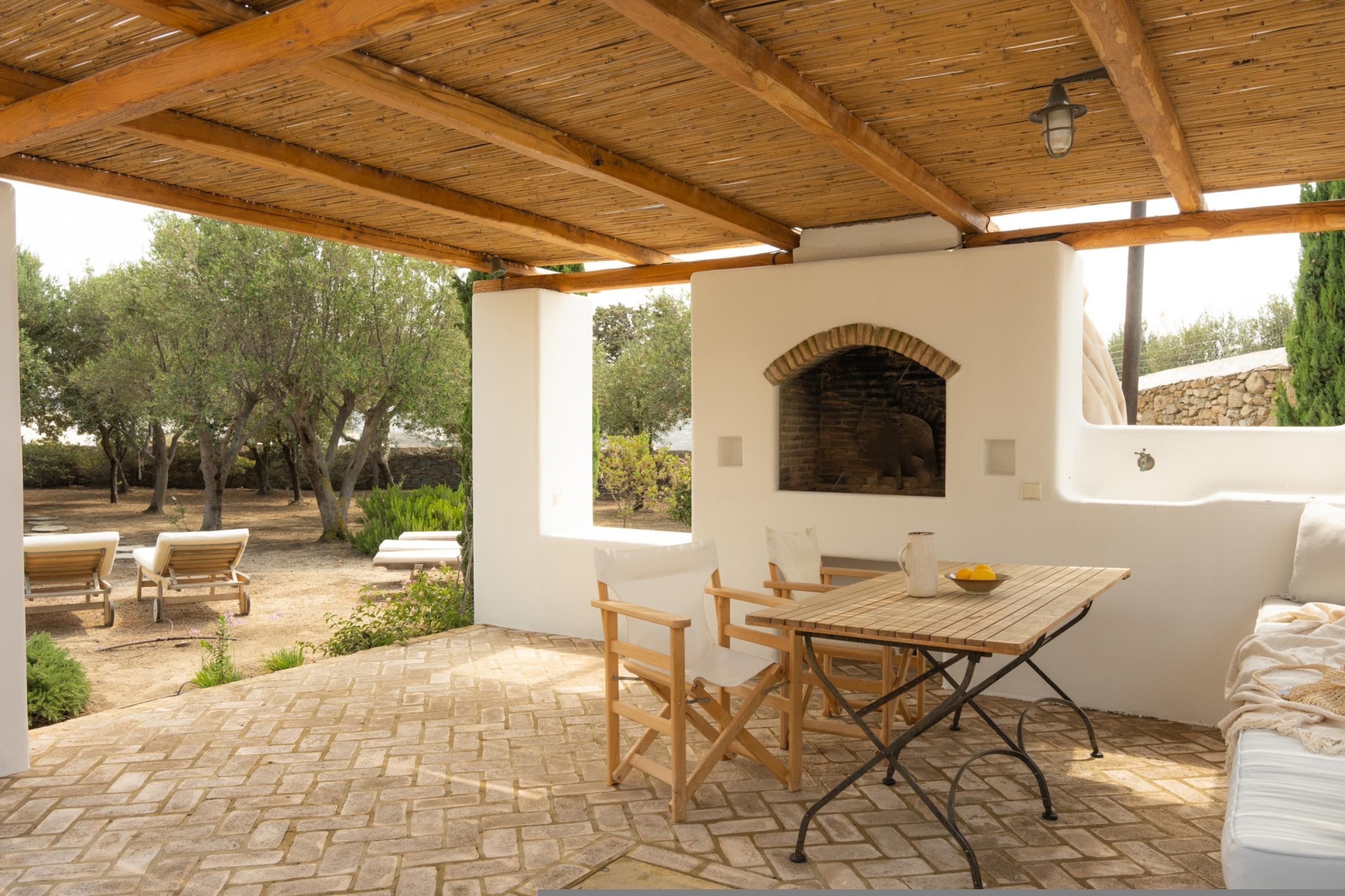Une terrasse extérieure avec un sol en briques, des meubles en bois et un mur en pierre agrémenté d’une cheminée en briques.
