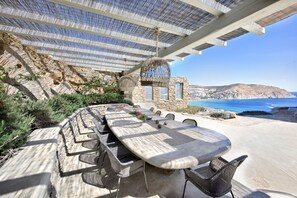 Zona d'exterior per dinar amb una taula de pedra i cadires de vimet, amb vista a un paisatge costaner i una visió clara del mar i les muntanyes llunyanes.