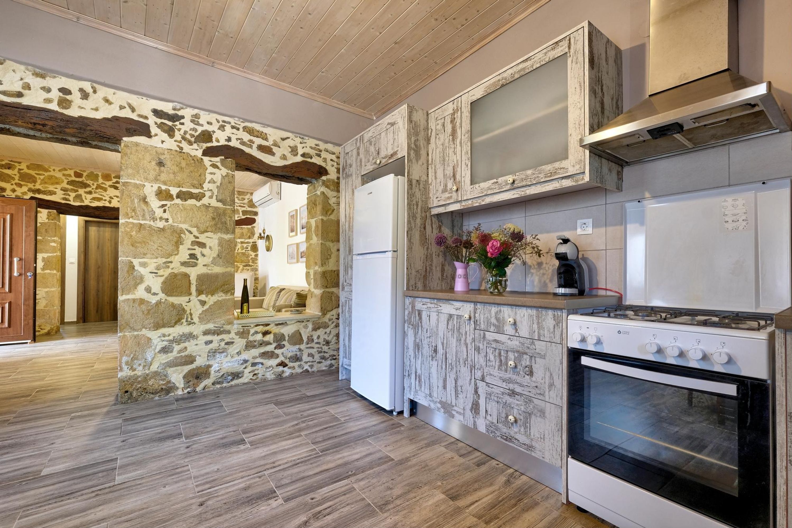 Una cocina con paredes de piedra, techo de madera, refrigerador blanco, estufa y un mueble de acabado rústico.