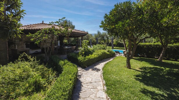 Un vialetto in pietra conduce a una casa in pietra con tetto a tegole, circondata da un verde lussureggiante e una piscina.